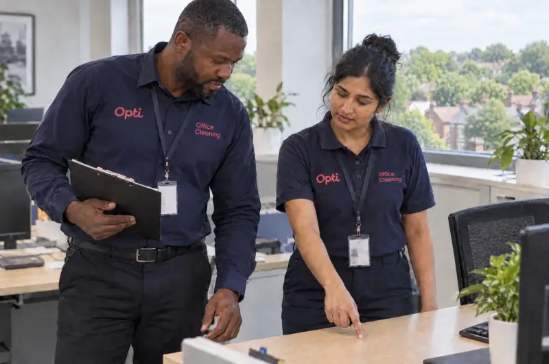 A supervisor in the navy button-up shirt checking a just-cleaned desk and washroom against a checklist, an operative alo
