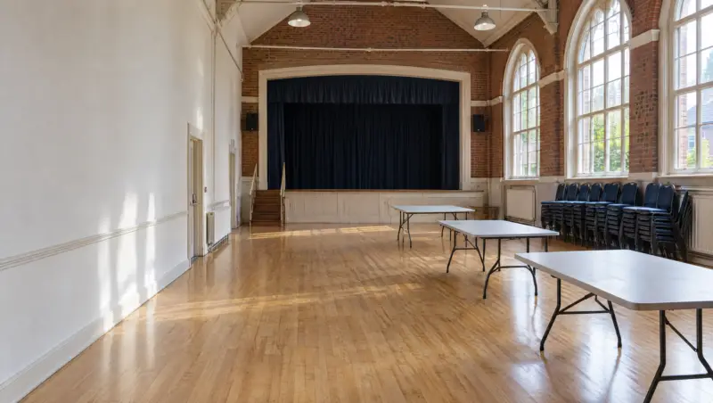 A clean community hall with a polished wooden floor, stage and stacked chairs